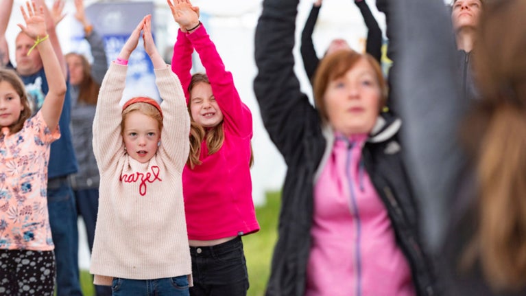 Group yoga workshop for families of all ages, a group of children practising yoga is in focus with the adults blurred out to the side.